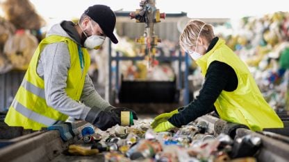 Two masked workers in high-visibility vests, sorting recyclables on a conveyor belt.